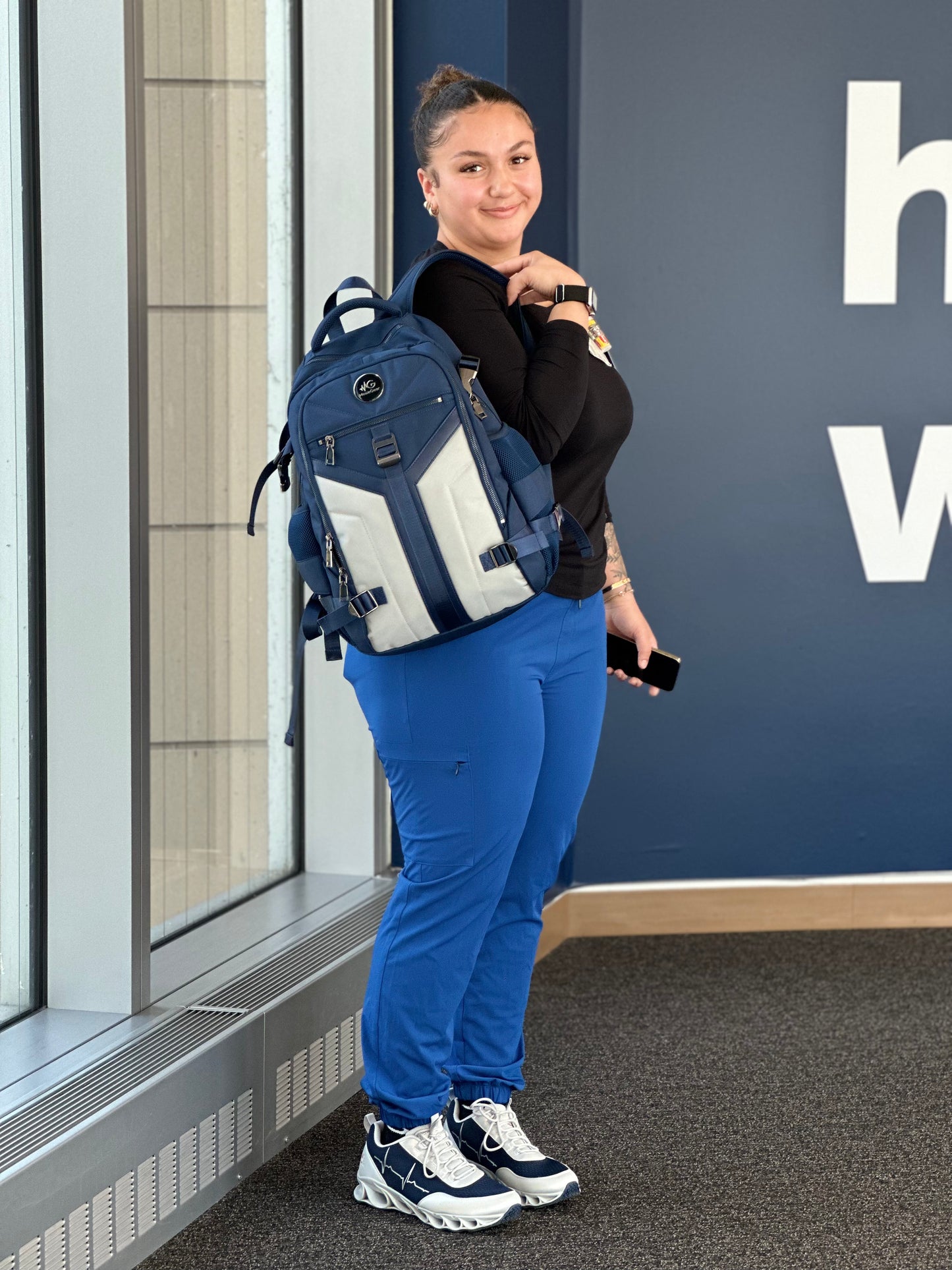 A nurse practitioner in moxiegalia premium scrubs standing indoors, wearing MG HoofForce IV sneakers and carrying the MG Aviator IJ backpack.