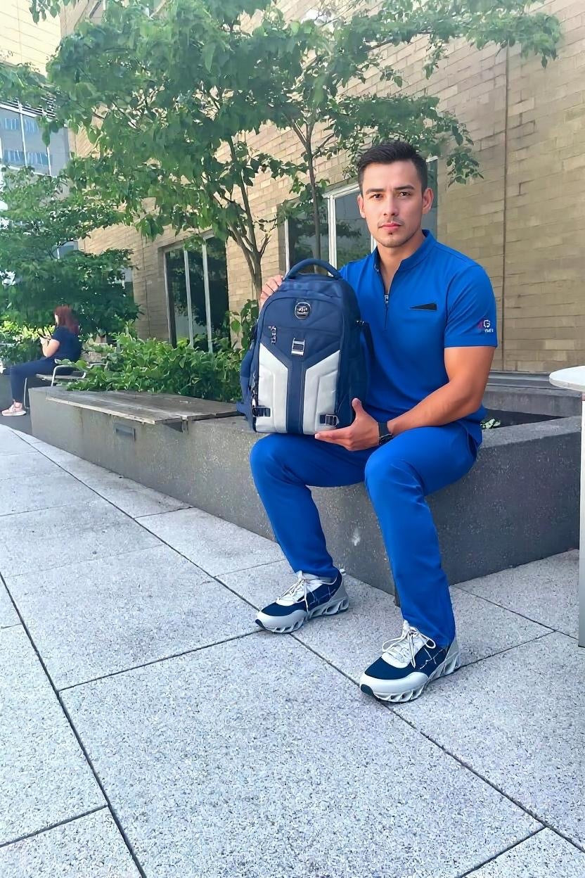 Man in blue scrubs sitting outdoors, holding the MG Aviator IJ backpack and wearing MG HoofForce IV sneakers.